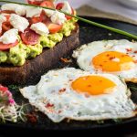 Appetizing avocado toast with cherry tomatoes and fried eggs, garnished with herbs.