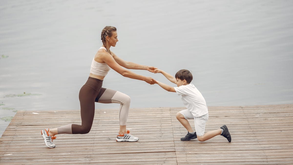 A woman and a boy exercise together on a wooden deck by the lake, promoting healthy lifestyle.